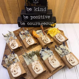 May include: Eight rectangular wax melts decorated with dried flowers, herbs, and fruit slices. Each melt is wrapped in a brown paper sleeve featuring a wooden tag shaped like a mason jar with a bee logo. Behind them are three stacked black signs.