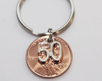 May include: A penny keychain with a silver number "50" charm attached. The penny is dated 1966 and is attached to a silver keyring. The penny is copper-colored, and the background is white.