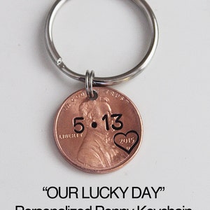 May include: A personalized penny keychain with a silver key ring. The penny has the date "2015" and the inscription "5.13" with a heart above the date. The text "OUR LUCKY DAY" is printed below the image.