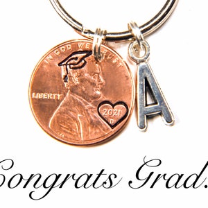 May include: A copper penny keychain with a graduation cap and heart with the year 2021 engraved on it. A silver charm with the letter A is attached to the keychain. The keychain is on a white background with the text "Congrats Grad!" below it.