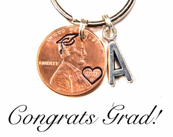 May include: A copper penny keychain with a graduation cap and heart with the year 2021 engraved on it. A silver charm with the letter A is attached to the keychain. The keychain is on a white background with the text "Congrats Grad!" below it.