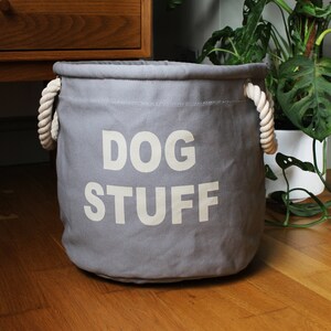 Peut inclure: Un bac de rangement en toile grise avec les mots "DOG STUFF" imprimés en blanc. Le bac a des poignées en corde et est conçu pour les fournitures pour animaux de compagnie. Le bac est sur un sol en bois.