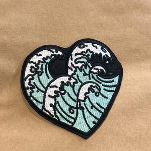 Puede incluir: Parche en forma de corazón con un diseño estilizado de olas oceánicas. El parche tiene un borde negro, con las olas representadas en tonos de verde azulado, blanco y negro. El diseño recuerda al arte japonés.