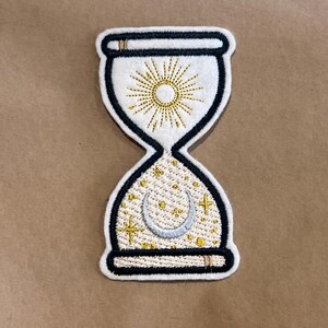 Könnte beinhalten: Gestickter Sanduhr-Aufnäher mit einem Himmelsdesign. Der Aufnäher hat einen weißen Hintergrund mit einem schwarzen Rand. Die obere Hälfte hat ein goldenes Sonnenmuster, während die untere Hälfte einen Mond, Sterne und Kreuze aufweist.