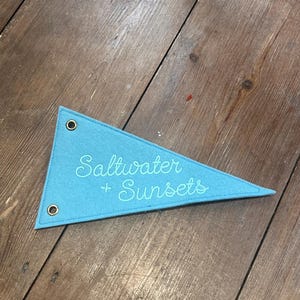 Może przedstawiać: Błękitny filcowy proporczyk z wyhaftowanym napisem "Saltwater + Sunsets". Proporczyk jest trójkątny z dwoma metalowymi oczkami i obszytą krawędzią. Proporczyk leży na drewnianej powierzchni.