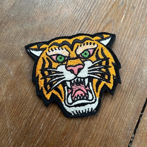 Può includere: Un primo piano di una toppa ricamata con il viso di una tigre. La tigre ha strisce arancioni e nere, occhi verdi e un naso rosa. La toppa è su uno sfondo di legno.