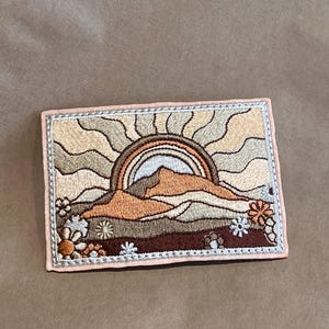 May include: Embroidered patch depicting a sunset over mountains, a rainbow, and flowers. The patch has a light pink border and is in shades of brown, orange, and beige. The design is detailed and textured.