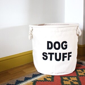 Peut inclure: Un panier de rangement en toile blanche avec des poignées en corde et le texte "DOG STUFF" imprimé en noir.