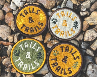 May include: Four embroidered patches with a brown border and yellow, green, and white stitching. Each patch says "Tails & Trails" with a paw print and a pine tree design.