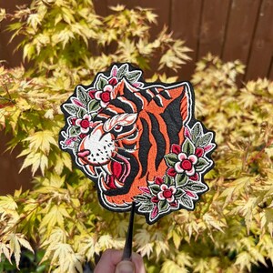 May include: An embroidered iron-on patch featuring a tiger's head with orange and black stripes. The tiger's head is surrounded by a floral design with red, pink, and green flowers.