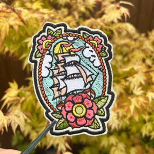 May include: Embroidered patch depicting a traditional sailing ship with white sails, set against a sky with clouds and sun. The design is framed by a rope border and floral accents in pink, yellow, and green. The patch has a black border.