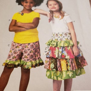 May include: Two girls wearing different skirts and tops. The girl on the left is wearing a yellow short-sleeved shirt and a pink and floral skirt with a ruffle trim. The girl on the right is wearing a white short-sleeved shirt and a multi-colored tiered skirt with a ruffle trim.