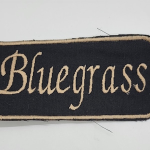 May include: A rectangular black patch with the word "Bluegrass" embroidered in a cursive, tan-colored font. The patch has a tan border and is likely designed to be sewn onto clothing or accessories.