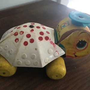 May include: A vintage wooden pull toy turtle with a white metal shell with red dots. The turtle has a yellow face with blue eyes and a blue spinning wheel on top. The toy is labeled "Tiny Tim".