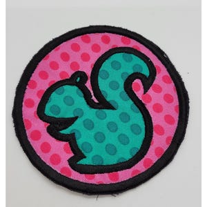 May include: A round patch featuring a teal squirrel design with black outlines, set against a pink background with red polka dots. The patch has a black border and is likely an iron-on or sew-on applique.