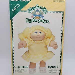 May include: A See & Sew 5423 Cabbage Patch Kids sewing pattern envelope featuring a doll with yellow yarn hair in pigtails, blue eyes, and a yellow dress with white trim. The text reads Cabbage Patch Kids, P'tits Bouts de Choux, and CLOTHES.