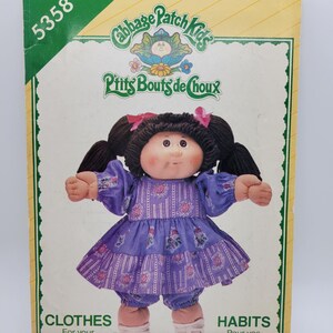 May include: A See & Sew 5358 sewing pattern envelope for Cabbage Patch Kids clothes. The package features a doll with black yarn hair in pigtails, wearing a purple floral dress and matching bloomers, with text including Cabbage Patch Kids.