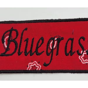 May include: A rectangular patch with the word "Bluegrass" embroidered in black script on a red background. The patch has a black border and a bandana-style pattern with white and red floral designs.