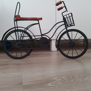 May include: A decorative metal bicycle model featuring a black frame, thin spoked wheels, a front wire basket, and a brown wooden seat. The bicycle includes a chain, pedals, and handlebars with brown wooden grips, designed as a stationary tabletop ornament.