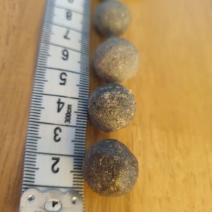 Può includere: Una serie di cinque piccole sfere testurizzate di colore grigio-marrone disposte in linea verticale accanto a un metro a nastro bianco. Il metro indica che le sfere hanno un diametro compreso tra circa 1,2 cm e 1,9 cm.