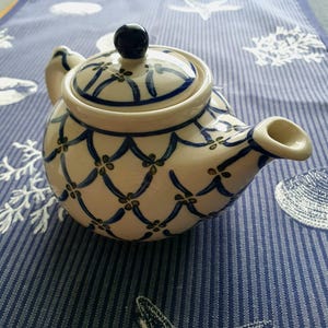 May include: A ceramic teapot with a cream-colored base featuring a blue lattice pattern and small green floral accents. The teapot includes a matching lid with a dark blue knob, a curved spout, and a handle, all decorated with the same pattern.
