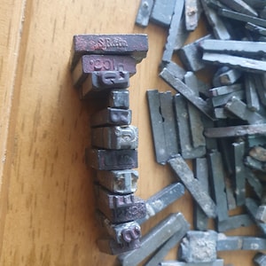 May include: A collection of rectangular metal letterpress type blocks, with one vertical stack showing characters including S, R, and Co, and the word pica. The individual pieces are dark grey, weathered, and metallic, with various letters and numbers embossed on their ends.