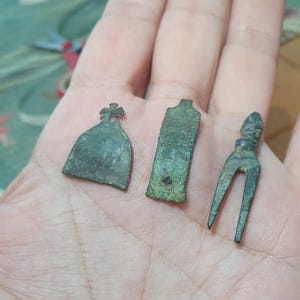 Puede incluir: Tres pequeños artefactos metálicos de tono bronce desgastado. Uno tiene forma de campana con una parte superior en cruz, otro es una placa rectangular plana con marcas texturizadas y el tercero es un objeto de dos puntas con mango.