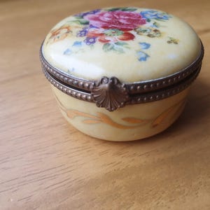 May include: A small, round, cream-coloured porcelain trinket box with a hinged lid. The lid features a painted floral design with pink, red, and blue flowers. The base is decorated with gold-toned leaf patterns and a bronze-coloured metal rim.