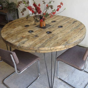 May include: A round wooden table with a rustic finish, supported by four hairpin legs. The table is made from reclaimed wood and has a natural wood grain. The table is set with two chairs with brown fabric seats.