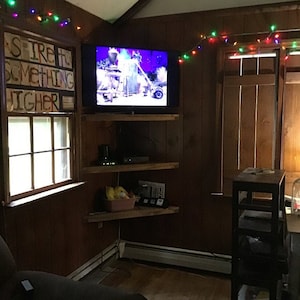 Puede incluir: Una habitaci&oacute;n acogedora con un televisor montado en estantes de madera. Se ve una ventana con el texto "STRE TH SOMETHING HIGHER". Luces de cadena est&aacute;n colgadas en la parte superior de la habitaci&oacute;n. Tambi&eacute;n hay una mesa peque&ntilde;a y una ventana con contraventanas de madera.