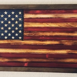 Op de afbeelding: Een Amerikaanse vlag van hout met een verweerde, verbrande uitstraling. De vlag is ingelijst in een donkerhouten lijst.
