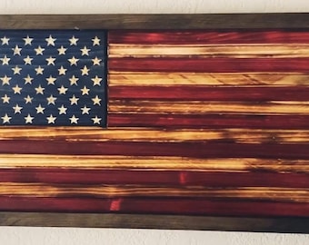 Puede incluir: Una bandera americana de madera con un aspecto desgastado y quemado. La bandera está enmarcada en un marco de madera oscura.