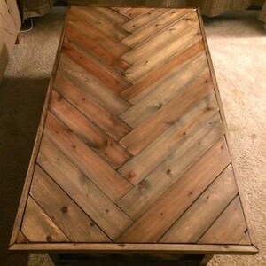 May include: Rectangular wooden coffee table with a herringbone pattern. The tabletop is constructed from planks of varying shades of brown and tan wood. The table has a dark brown frame and legs.