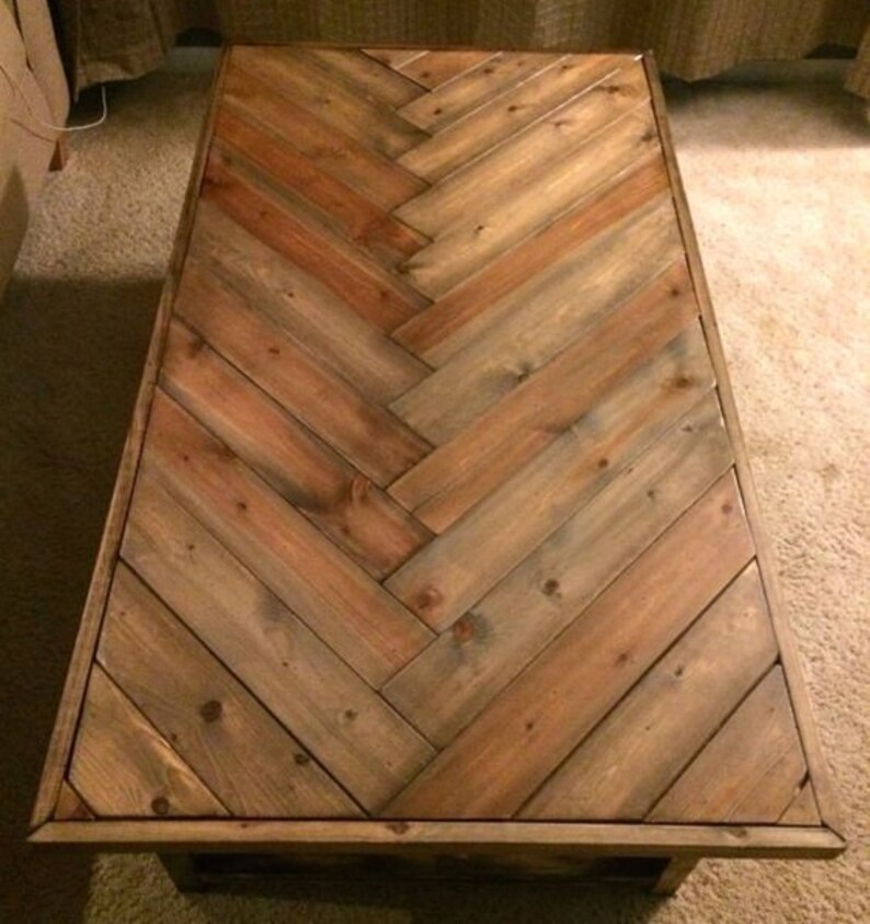 May include: Rectangular wooden coffee table with a herringbone pattern. The tabletop is constructed from planks of varying shades of brown and tan wood. The table has a dark brown frame and legs.