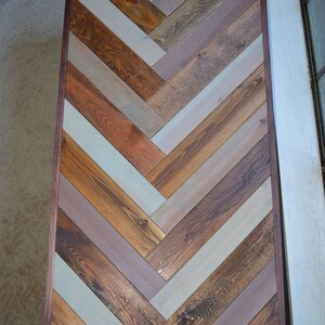 May include: Rectangular wooden table with a herringbone pattern. The table features a variety of wood tones, including light blue, brown, and orange, arranged in a chevron design. The table is framed with a dark brown border.