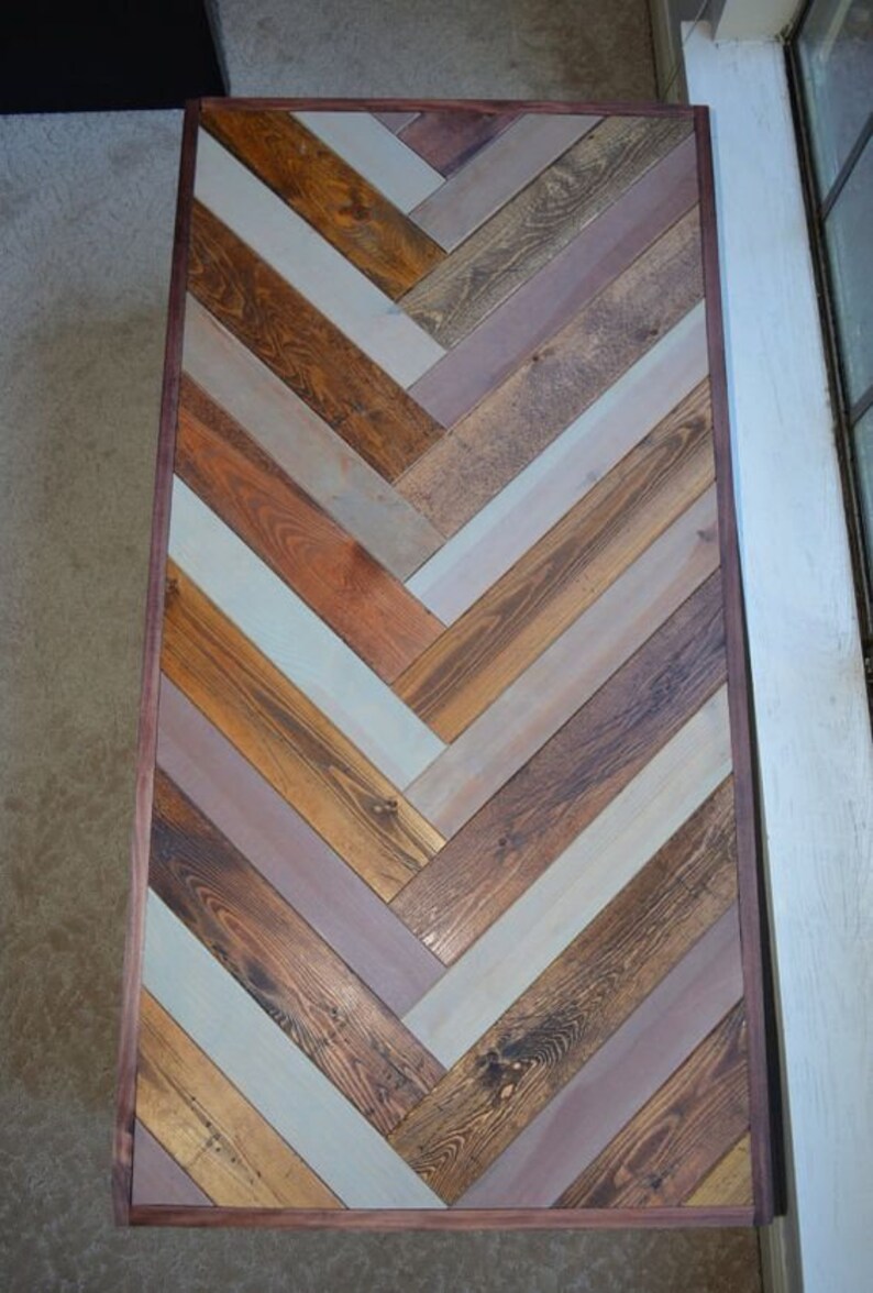 May include: Rectangular wooden table with a herringbone pattern. The table features a variety of wood tones, including light blue, brown, and orange, arranged in a chevron design. The table is framed with a dark brown border.