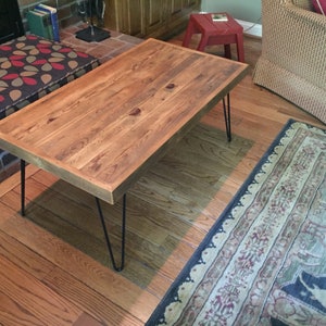 May include: A rectangular wooden coffee table with a natural finish. The table has a thick, solid wood top and is supported by four black hairpin legs. The table is approximately 48 inches long and 24 inches wide.