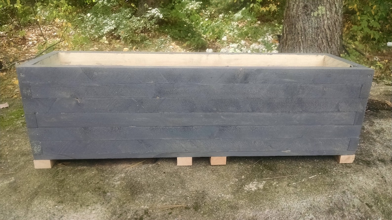 Op de afbeelding: Een grote, rechthoekige plantenbak gemaakt van donkergrijs hout met een natuurlijke houten bovenkant. De plantenbak staat op vier kleine houten blokken.