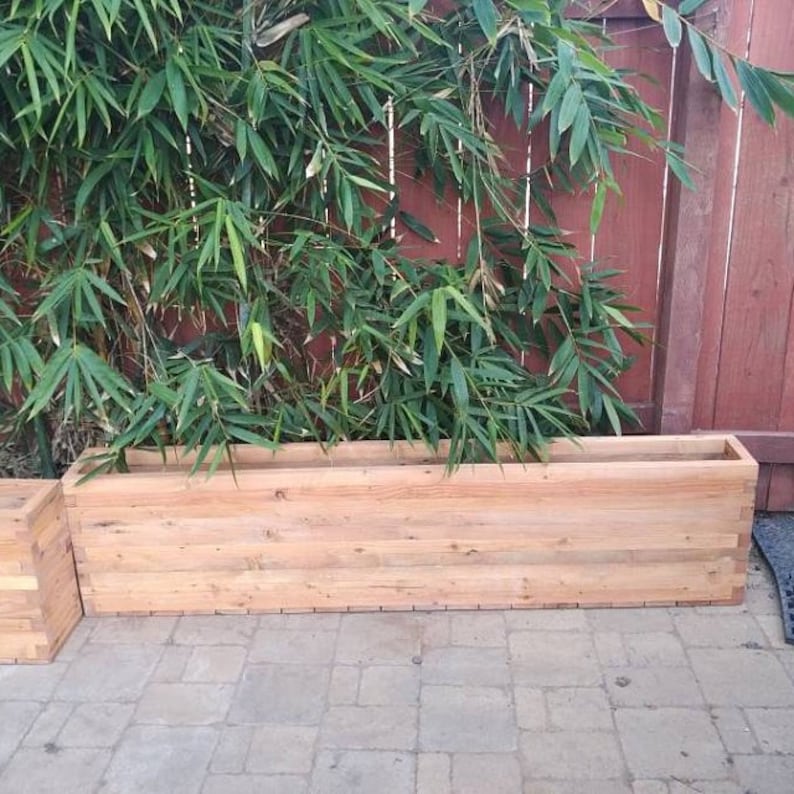 Op de afbeelding: Een lange, rechthoekige houten plantenbak gemaakt van natuurlijke houten planken. De plantenbak is leeg en staat op een terras van bakstenen.