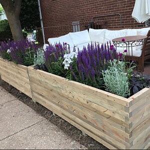 Op de afbeelding: Twee lange houten plantenbakken gevuld met paarse en witte bloemen. De bakken zijn gemaakt van gerecycled hout en hebben een rustieke uitstraling.