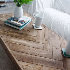 May include: A rustic, wooden coffee table with a herringbone pattern. A white vase with green leaves, two books, and a white mug on a coaster are on the table. The table is next to a white sofa.