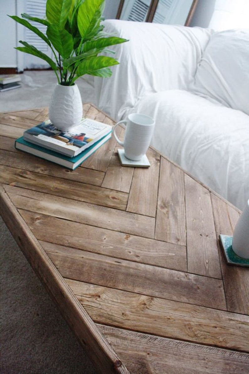 May include: A rustic, wooden coffee table with a herringbone pattern. A white vase with green leaves, two books, and a white mug on a coaster are on the table. The table is next to a white sofa.