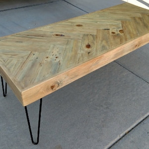 May include: A rectangular coffee table with a light wood herringbone pattern top. The table has a natural finish and is supported by four black hairpin legs. The table measures approximately 48 inches long.