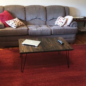 May include: A dark brown wooden coffee table with black hairpin legs. A silver laptop and a remote control sit on the table. The table is on a red rug in front of a gray sofa with decorative pillows.