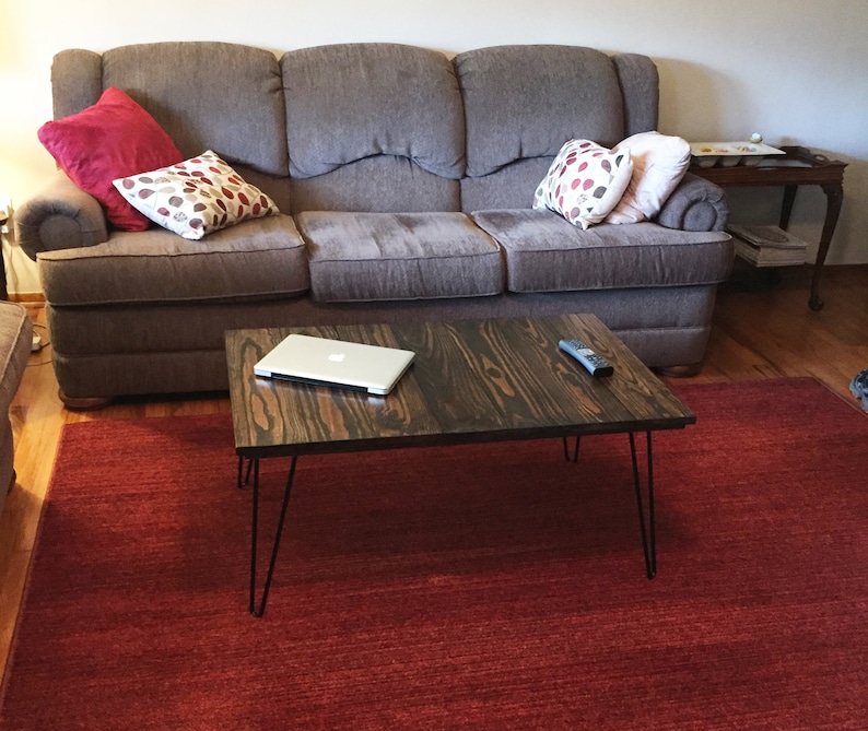 May include: A dark brown wooden coffee table with black hairpin legs. A silver laptop and a remote control sit on the table. The table is on a red rug in front of a gray sofa with decorative pillows.