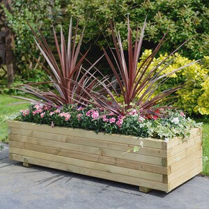 Op de afbeelding: Een houten plantenbak met een natuurlijke afwerking, gevuld met roze bloemen en roodbladige planten. De plantenbak staat op kleine pootjes en staat op een grijs terras.