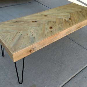 May include: A rectangular wooden coffee table with a light-colored, herringbone-patterned top. The table has a natural wood finish and is supported by four black hairpin legs. The table measures approximately 48 inches long.