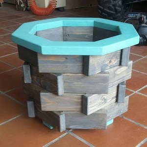 May include: Octagonal wooden planter with a teal blue rim. The planter is made of stacked wooden planks in a geometric pattern. The planter is sitting on a red tile floor.