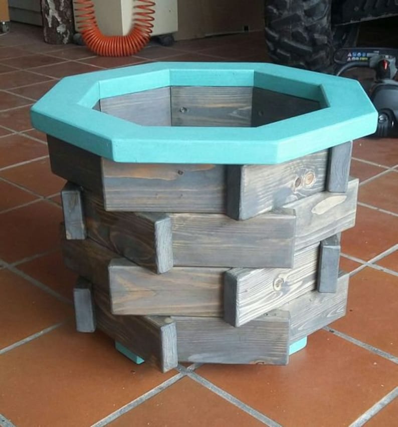 May include: Octagonal wooden planter with a teal blue rim. The planter is made of stacked wooden planks in a geometric pattern. The planter is sitting on a red tile floor.