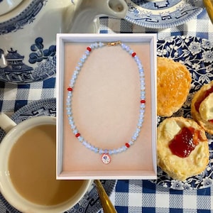 Könnte beinhalten: Eine Perlenkette mit hellblauen facettierten Perlen, kleinen goldfarbenen Zwischenelementen und leuchtend roten Perlen in regelmäßigen Abständen. Die Kette hat einen zentralen ovalen Anhänger mit weißem Hintergrund und einer kleinen violetten Blumenillustration.
